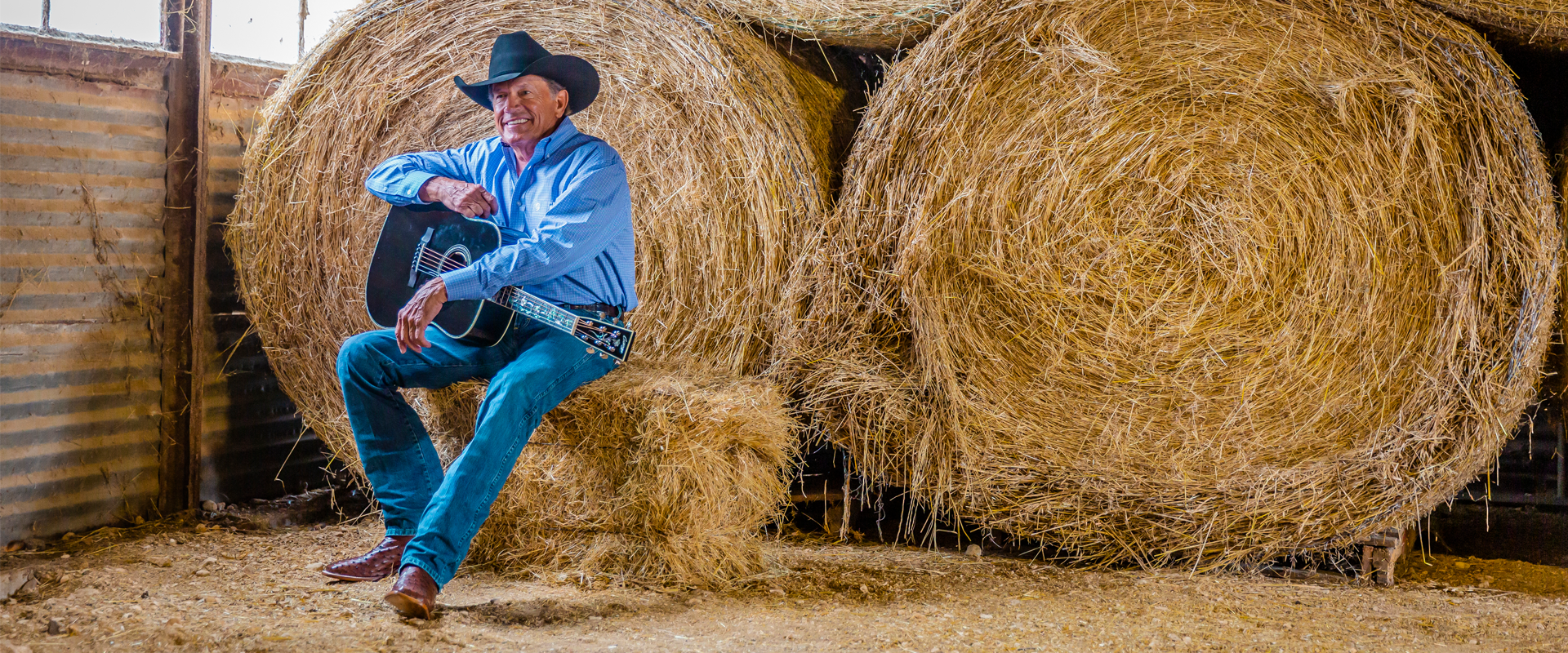 George Strait sitting on a hay bail holding a guitar.